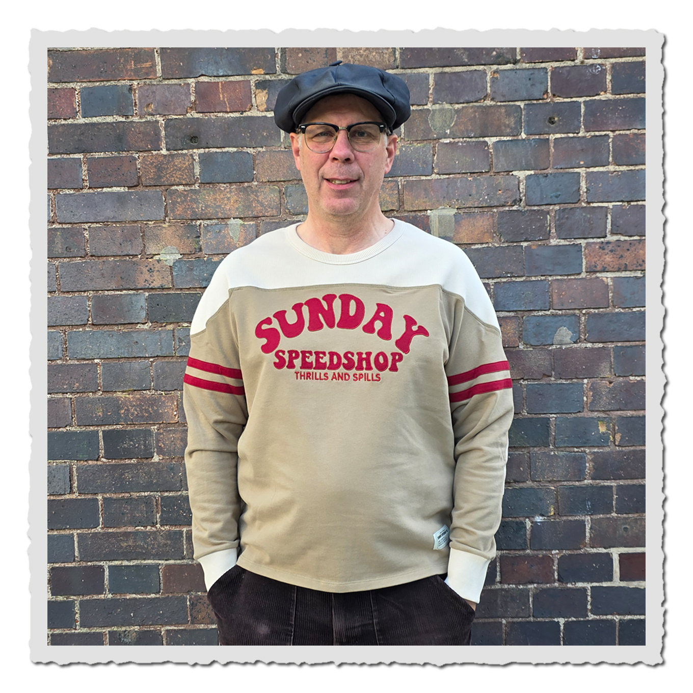 Mann trägt beiges Langarmshirt mit hellem Schulterbereich und rotem Frontmotiv vor Backsteinwand.